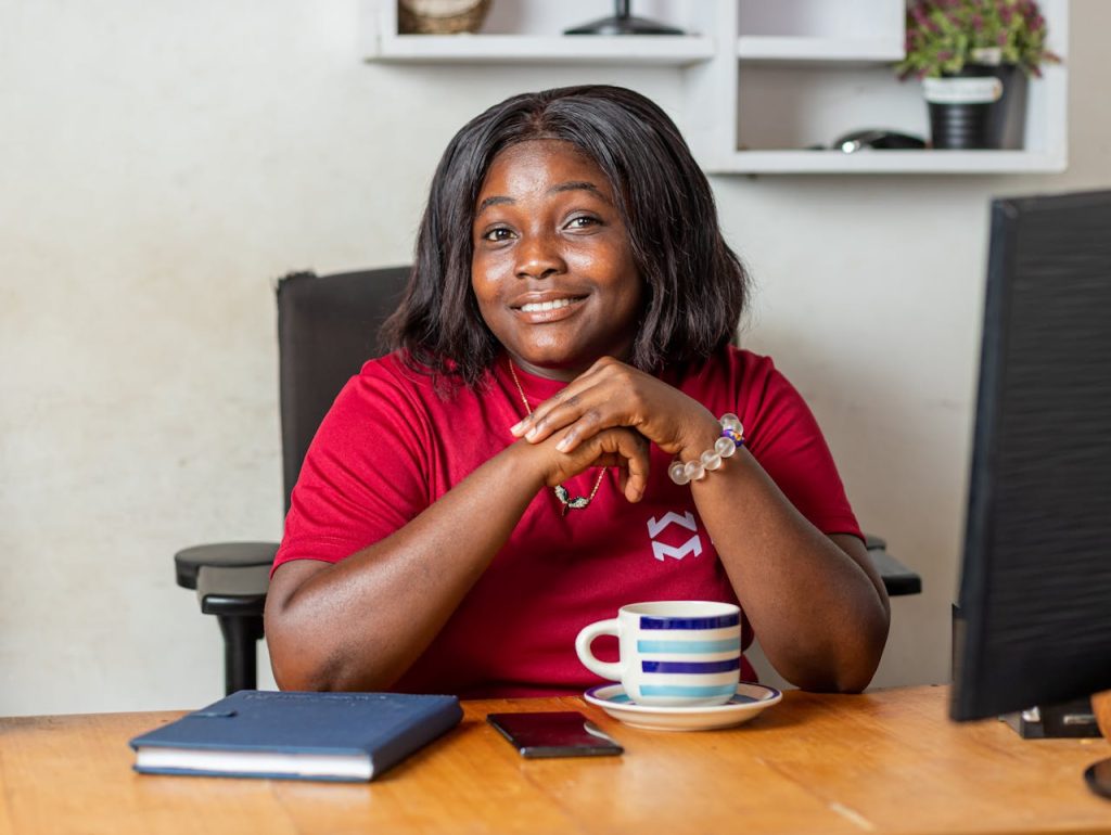 Portrait of a smiling African American woman sitting at a desk in an office setting with coffee and notebook.